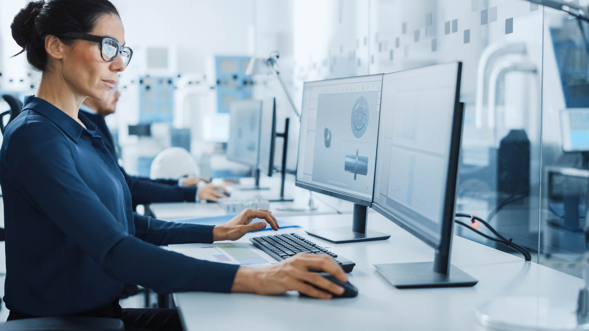 Ingenieurin mit Brille arbeitet an CAD-3D-Modellen auf zwei Monitoren in einem modernen Technikbüro