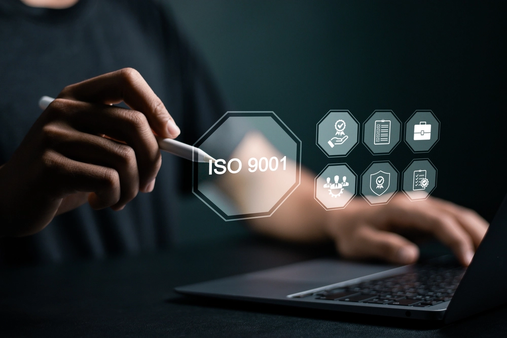 Person zeigt mit Stift auf ISO-9001-Symbol mit digitalen Qualitätsmanagement-Icons über einem Laptop
