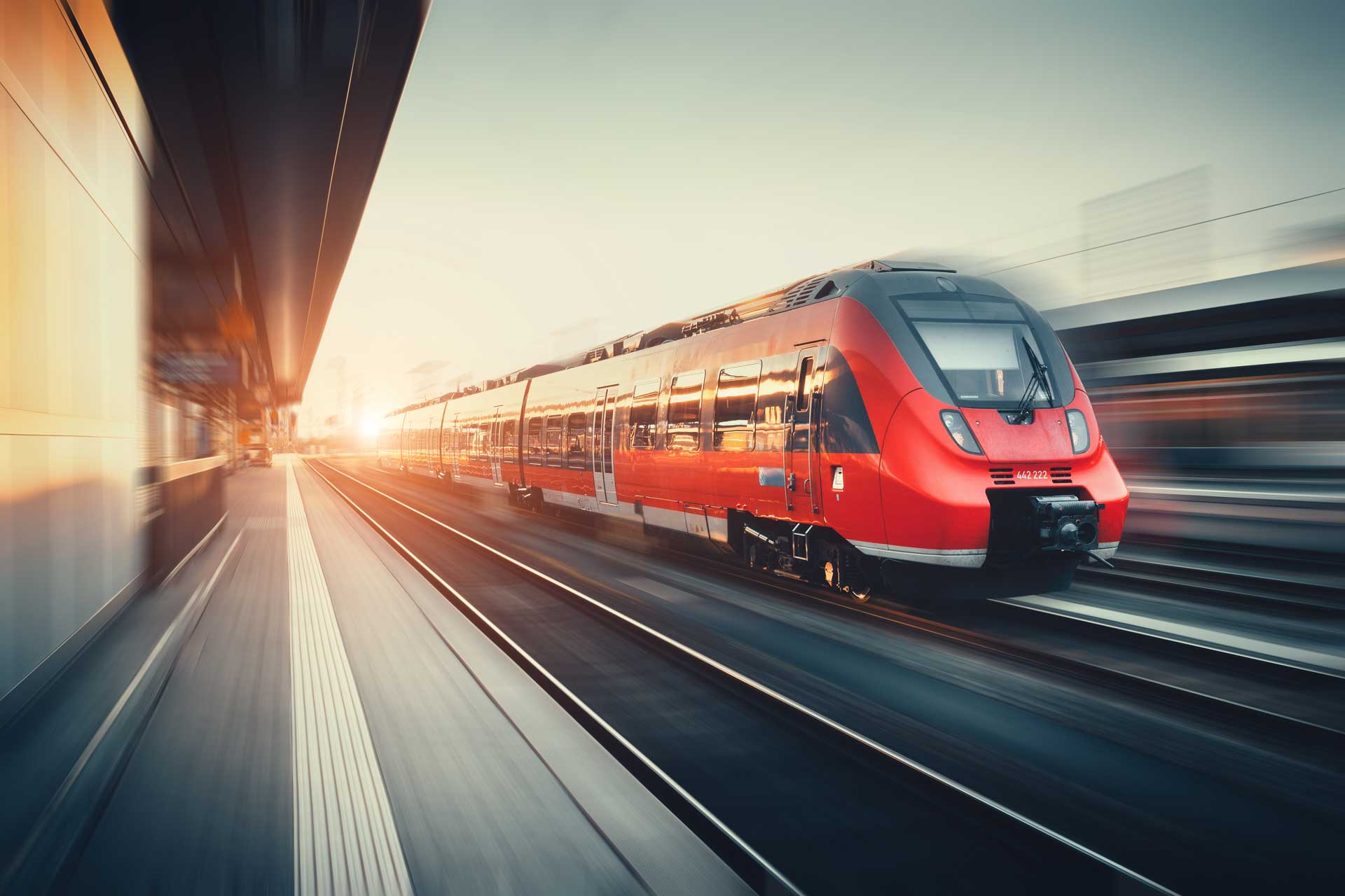 Roter Hochgeschwindigkeitszug durchfährt mit Bewegungsunschärfe einen Bahnhof bei Sonnenuntergang