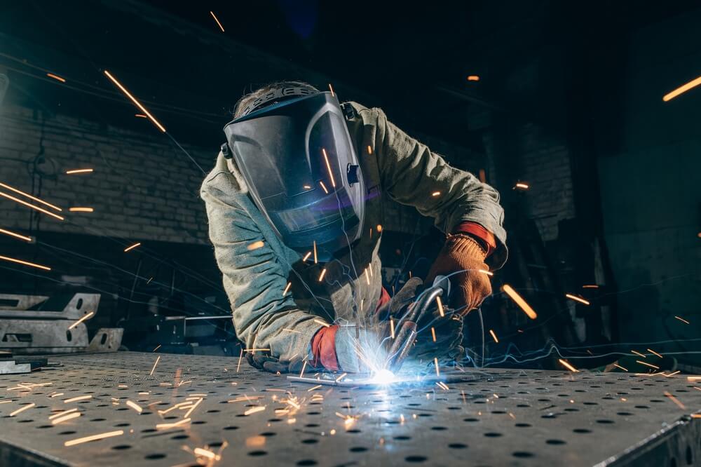 Person mit Schweißhelm und Handschuhen bei Schweißarbeiten an einer Metallplatte, Funken sprühen in der Werkhalle.