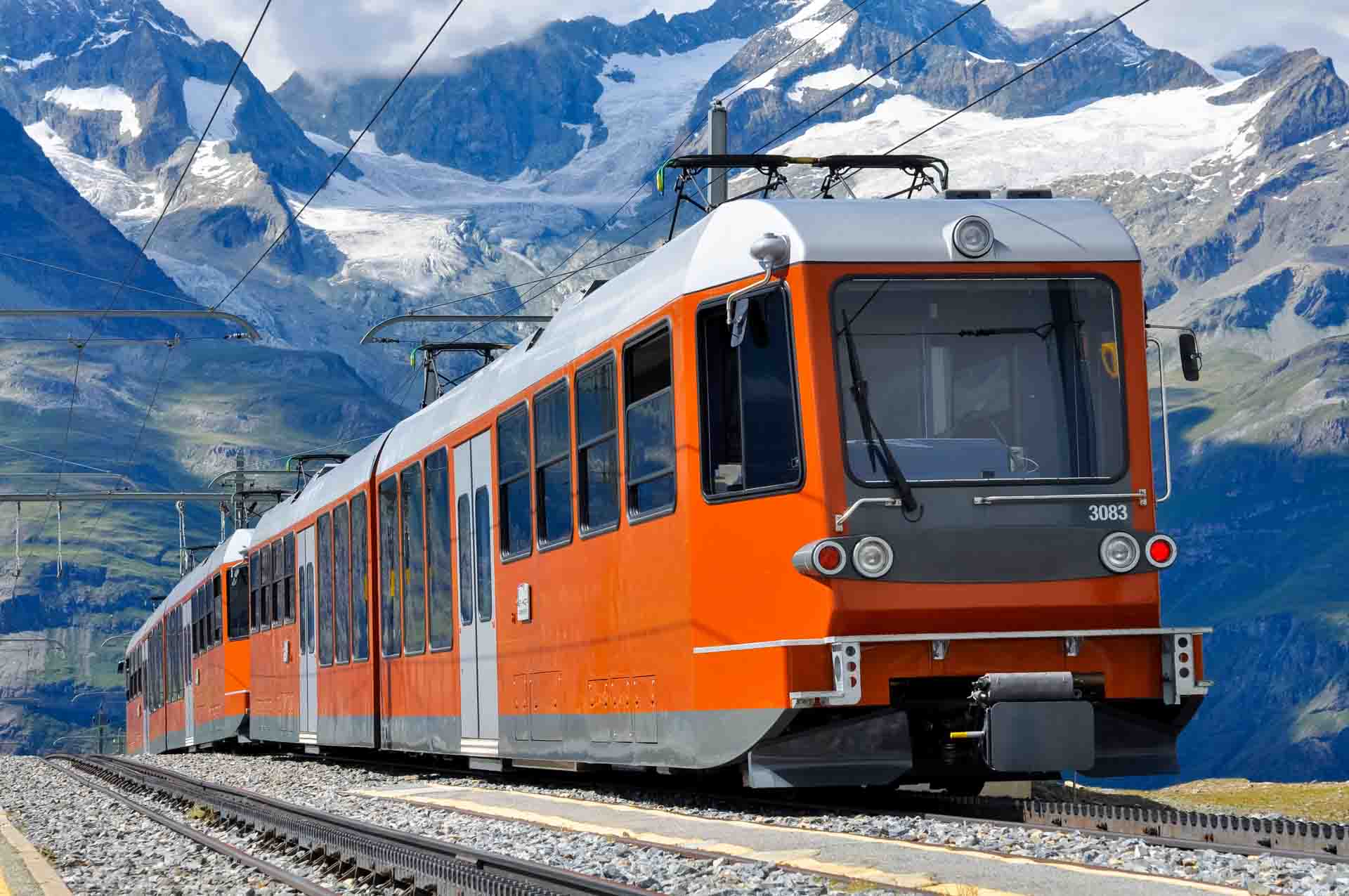 Orangefarbener Bergbahn-Triebwagen 3083 fährt vor schneebedeckten Alpengipfeln auf einer Zahnradbahn
